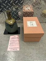 Japanese ceramic vase with green and brown glaze, black base, tan box, and Japanese information sheet on gray metallic surface.