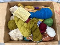 Large box filled with assorted yarn skeins and balls in green, blue, cream, and other colors, with some wooden yarn holders.