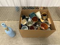 Overall view of cardboard box filled with large ceramic cups in assorted shapes, sizes, and colors, next to a wrapped bottle-shaped item.