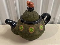 Side view of large ceramic teapot with textured green surface, swirl patterns in green and peach, black handle and spout, and ornamental lid.
