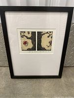 Full view of the framed art print leaning against a metal wall; shows black frame, white matting, and the artwork centered in the frame.