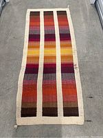 Top-down view of a vintage woven textile runner with three vertical stripes in warm colors, lying flat on a concrete floor.