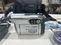 Front view of silver Hitachi DVD camcorder on white surface, showing Cinema Res branding and model details. Carry case visible in background.