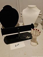Black velvet necklace bust, white necklace bust, black velvet bracelet stand with Fendi branding, and white ceramic hand-shaped ring holder with red painted nails photographed on table.