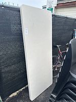 Side view of the folded Lifetime folding table resting vertically against a fence outdoors, showing beige plastic top with some dirt marks.