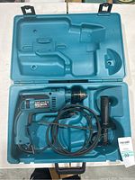 Open teal plastic case showing the Makita hammer drill with attached cord and the auxiliary handle in its molded spot.