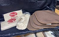 Four brown fabric seat pads and multiple new canvas tote bags with Utrecht logo, some bags still sealed in plastic packaging, all placed together on dark fabric surface.