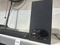 Set of Polk soundbar and subwoofer on a metal shelf. Side view of subwoofer shows black fabric covering and front circular bass port with Polk logo.