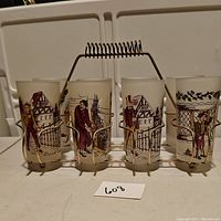 Front view of set showing all 8 Hazel Atlas frosted Charles Dickens character glasses in gold metal caddy with coil spring handle. Characters and vintage house imagery shown on each glass.