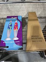 Two boxes on a metal shelf; one box is a Jambo double pack of lava lamps with right side of the lava lamp box visible, the other is a plain brown cardboard box labeled 'Ukulele'