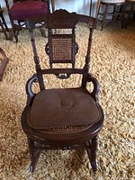 Front view of antique wooden rocking chair with brown cushioned seat and wicker backrest on carpet.