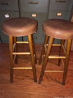 Pair of wooden bar stools with brown padded round seats on a wooden floor.