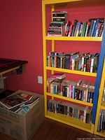 Yellow bookshelf with about 30 books arranged on four shelves, showing variety in sizes, colors, and thicknesses. Box with additional books to the side.