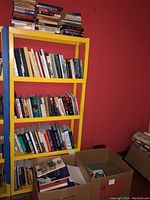 Photo showing a yellow bookshelf filled with various hardcover and softcover books, with a box of additional books on the floor nearby.