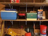 Wide view of wooden shelves showing the blue vintage Coleman cooler, red portable charcoal grill, green Coleman cooler, Stinger insect killer box, yellow and green garden rakes, garden shovels, and a large red bucket labeled 'Garage Bucket'.