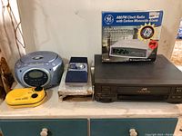 Lot of five electronic devices including a boxed GE AM/FM clock radio with carbon monoxide alarm, TMX boom box, JVC VCR, Hazor portable radio and yellow sport radio.