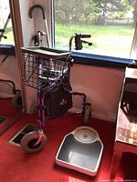 Purple Nova walker with black seat and wire basket, standing near window with red carpet background, also showing the bathroom scale on floor