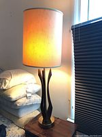 Single Mid-Century Modern table lamp illuminated on a side table, showing tall cylindrical fabric shade and sculptural wooden base with brass round platform.