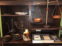 Photo showing a vintage brass scale, brass coffee urn, several plates, brass candleholder, and bird figurines arranged on dark wood shelves.