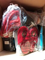 Box filled with multiple pairs of new slippers in packaging showing red, black, and blue colors with some holiday-themed prints