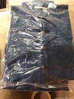Top view of a folded blue denim jacket in plastic packaging, showing metal buttons and front pockets.