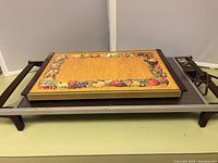Side view of one warming tray showing fruit border design on top and wooden handles