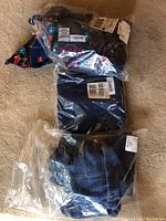 Three packaged women’s denim garments on floor: one shirt with floral embroidery, one pair of dark denim pants, one denim jacket with buttons.