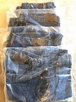 Four pairs of folded blue denim pants in plastic packaging showing leather patches on the waistband.