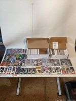 Two open boxes filled with baseball cards, additional cards displayed spread out on table surface. Cards cover various players and years.