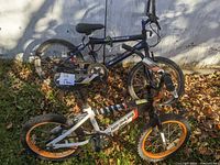 Two kids bikes leaning against a wall on dried leaves, showing both bikes' full profile and condition