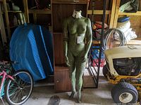 Front view of green painted female mannequin standing, showing whole figure with peeling paint and cracked texture.