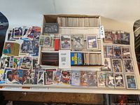 Wide view of the large box filled with thousands of assorted baseball cards, with a selection of cards laid out on the table around the box. Visible players include Deion Sanders and others. The box holds many unsorted cards in rows alongside some card packs and visible brand boxes.