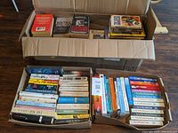 Three boxes filled mostly with paperback fiction books placed on the floor. Books arranged vertically and horizontally showing various titles and authors.