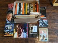 Photo showing assorted books including a cardboard box filled with old fiction and historical books, several music books like 'Led Zeppelin', 'Rock', and 'Baghdad or Bust',' and other notable hardcover novels.