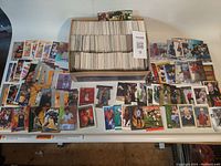 Photo showing a large shoebox filled with hundreds of assorted sports cards, surrounded by sample cards laid out on a table including basketball, football, hockey, and golf cards.