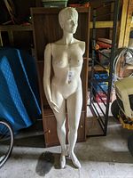 Full view of white female mannequin standing, visible one removable arm, located in storage area.