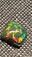Close-up image of the cushion-shaped Ethiopian black opal cabochon showcasing vibrant green, red, and orange flashes with a dark body tone on textured background.