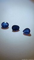 Three blue sapphire gemstones displayed on a white background, showing different angles and cuts of the stones in natural lighting.