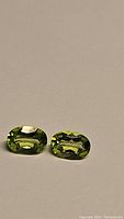 Two oval cut natural green peridot gemstones lying on a neutral background, showing their bright apple green color and facets.