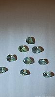 Eight natural Alexandrite gemstones arranged on a white background showing their pear and oval cuts and green to soft green-yellow color tones.