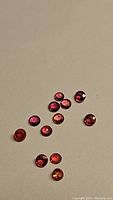 Twelve small round faceted sapphires with vivid fire red to orange color, scattered on plain surface, showing size and color consistency.