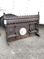Front view of dark stained antique pump organ top featuring detailed finials, carved trim, round mirror center, two small shelves, and wooden medallions on either side