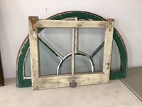 Green painted wooden arch window with glass, showing wear and chipped paint, and a smaller white weathered rectangular window with glass in front