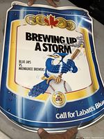 Blue Jays poster showing 'Brewing Up A Storm' with Blue Jays vs Milwaukee Brewers matchup and mascot holding baseball bat on a field colored background.