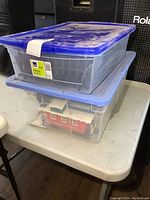 Two stacked transparent plastic totes with blue lids, one showing a red building model inside, likely related to the train set.