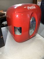 Red retro style Coca-Cola branded mini cooler fridge with rounded edges and chrome bottle opener on the side, placed on a table.