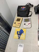 Two Nintendo Game Boys, one yellow heavily worn and one grey, placed on a table with a Super Mario Galaxy soundtrack booklet beneath them, and a Nintendo carry case above.