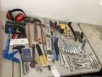 Wide view showing assortment of hand tools on a table including hammers, wrenches, screwdrivers, ear muffs, socket sets, and other small tools