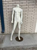 Full body view of the vintage white resin male mannequin standing upright on a round rusted metal base against a brick wall background.