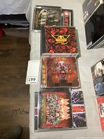 Top view of four music CDs stacked on bed showing Queen Greatest Hits, Aerosmith, Kiss, and Def Leppard albums.